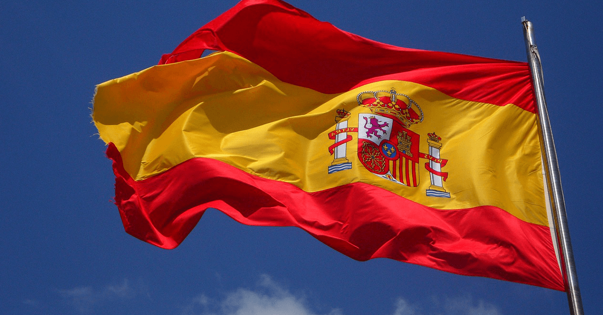 spain-flag A red-yellow-red Spanish flag with a coat of arms, flying in the wind against a blue sky.